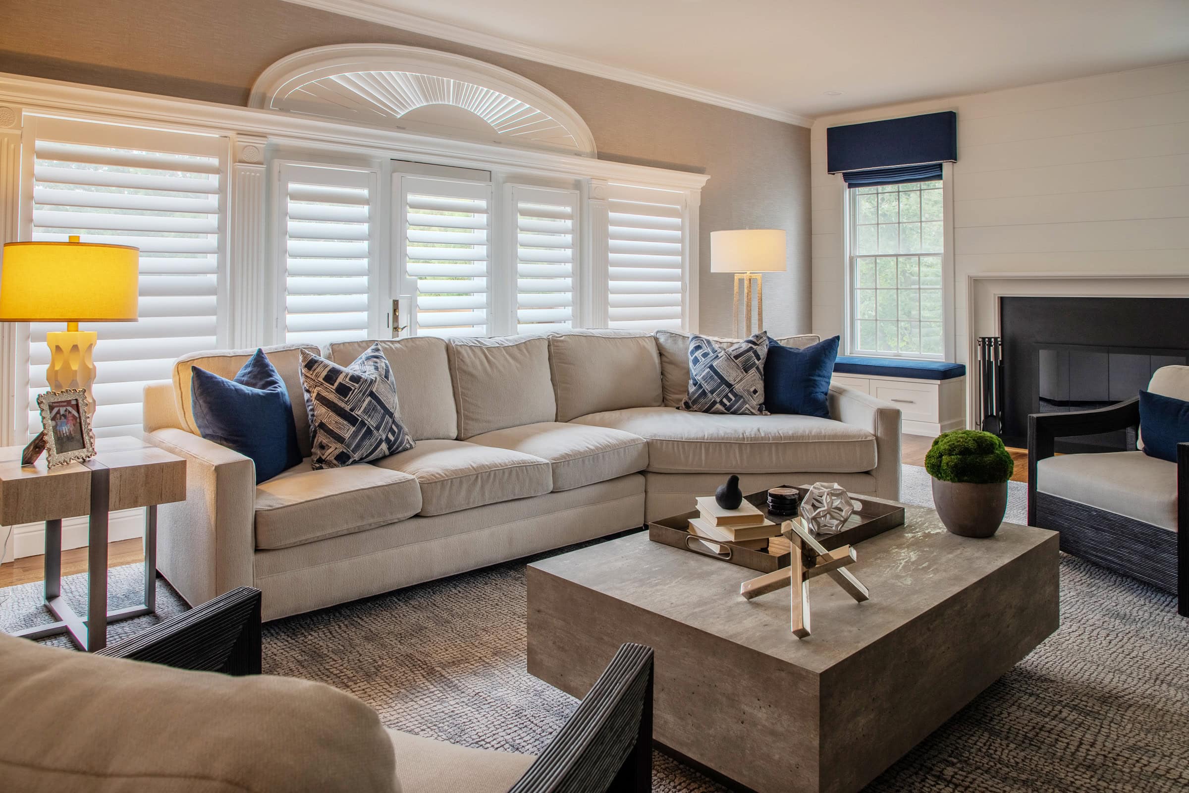 A modern living room with a beige sofa, blue and patterned pillows, a large stone coffee table, two side tables with lamps, window shutters, and a window seat with a blue cushion.