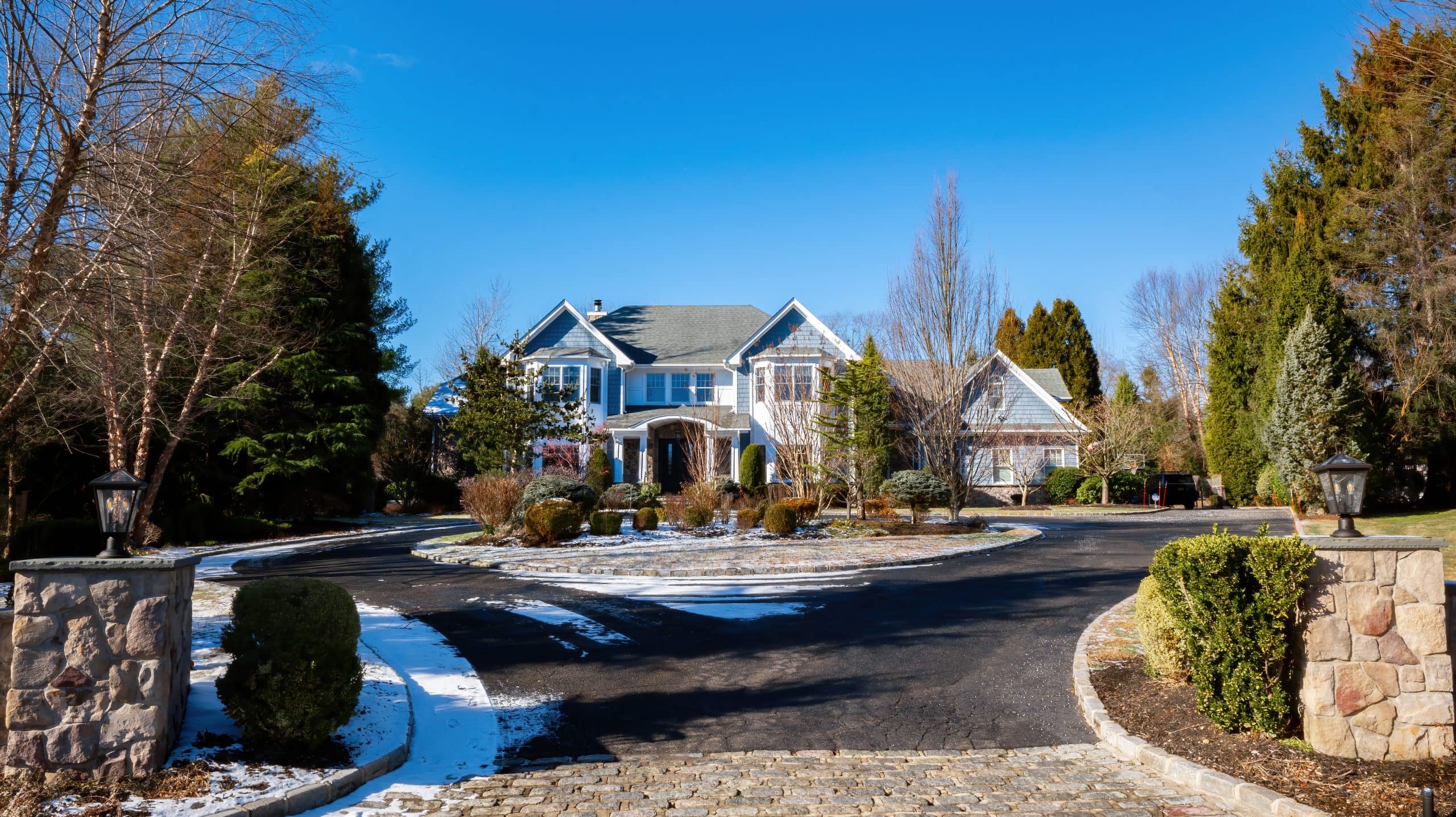 A large two-story house with a circular driveway and landscaped yard, surrounded by trees and patches of snow on the ground, under a clear blue sky. Stone pillars with lanterns mark the entrance.