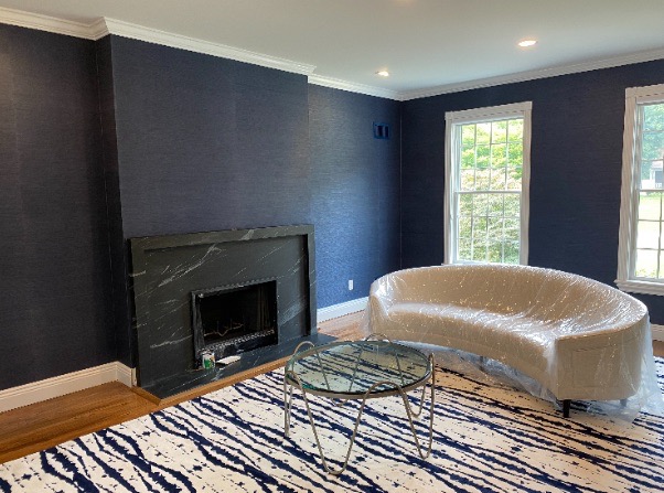 Living room mid-installation with navy grasscloth wallpaper, a black stone mantel, and a plastic-wrapped curved sofa