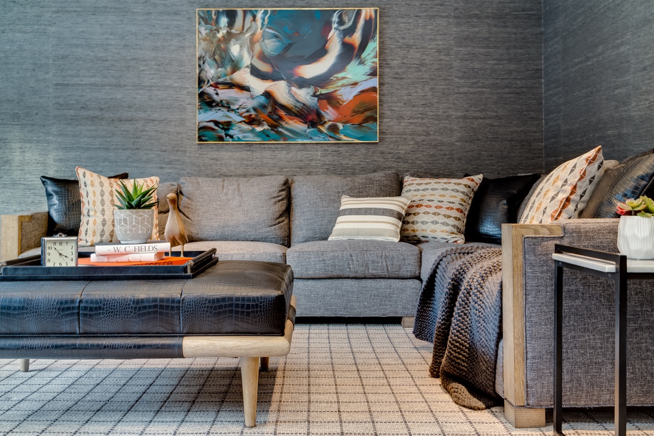 Den with grey grasscloth walls, charcoal sectional, leather ottoman, and a colorful abstract painting