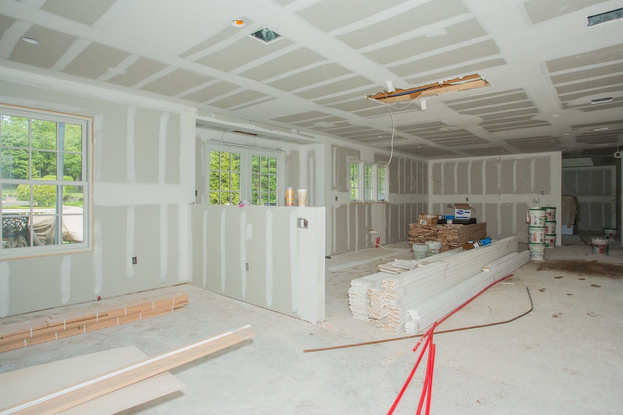 Kitchen under construction with drywall, framed window openings, and bundled trim and flooring on the subfloor