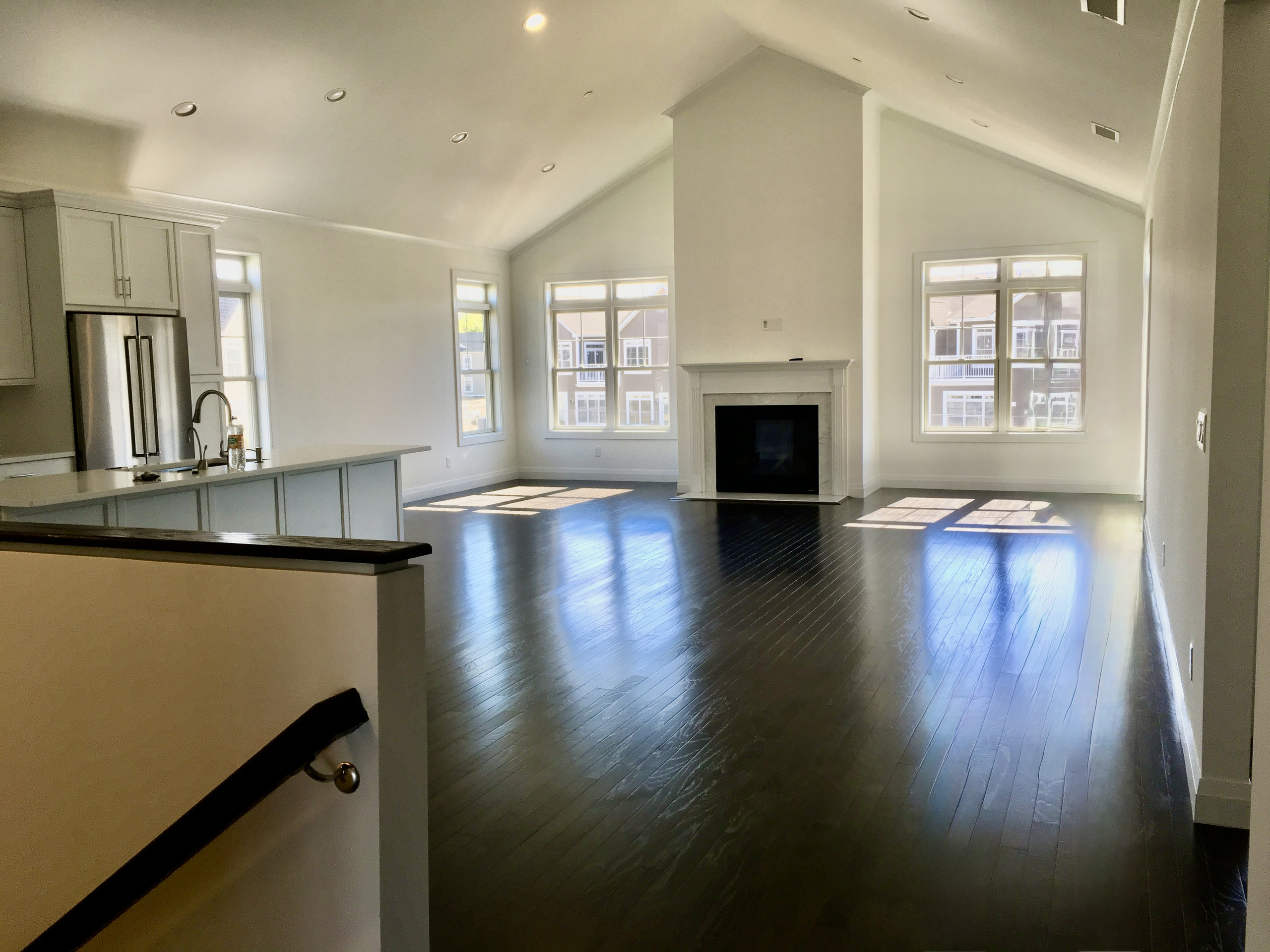 Empty great room before design with vaulted ceiling, plain builder fireplace mantel, and bare dark hardwood floors