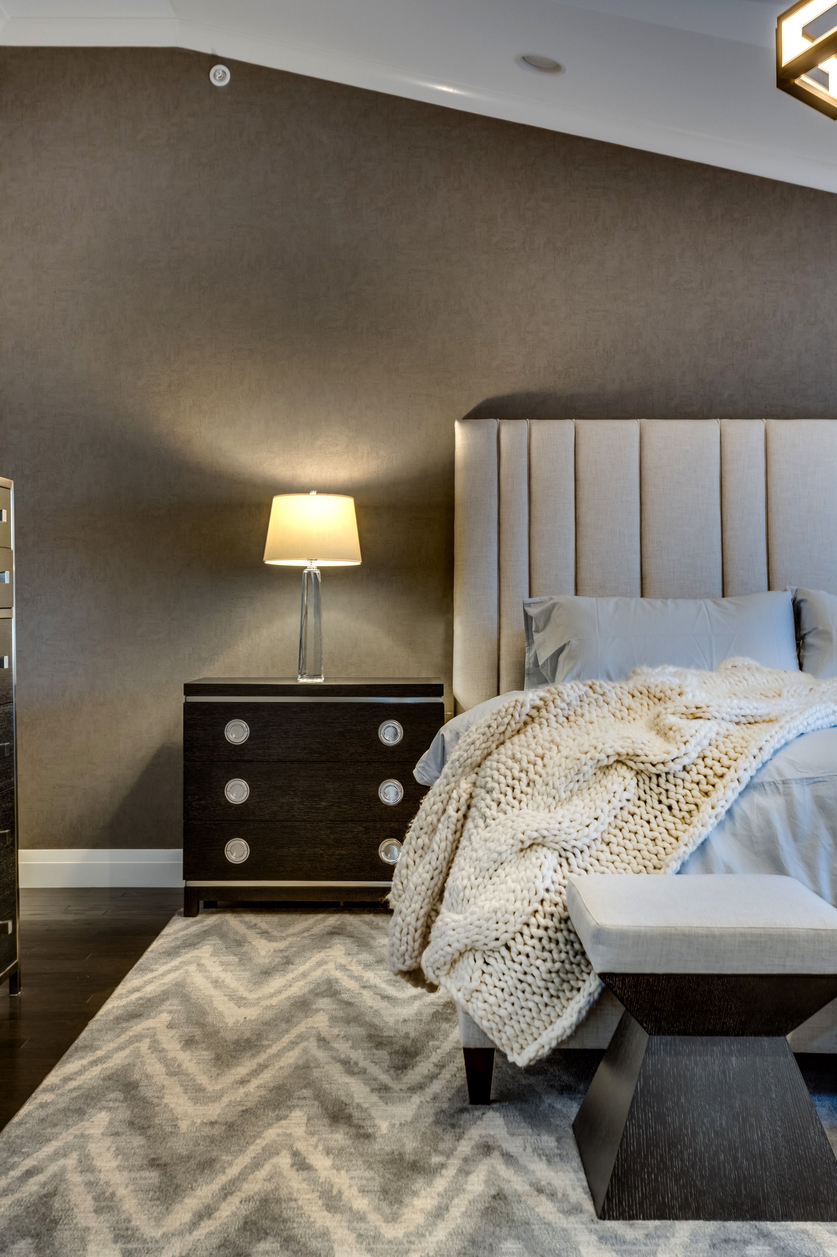 Bedside detail with dark wood nightstand, polished nickel ring pulls, crystal lamp, and chunky cream knit throw