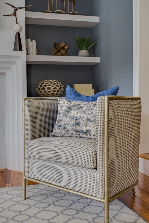 Woven barrel accent chair with brass bamboo-style frame and blue patterned pillows beside white floating shelves