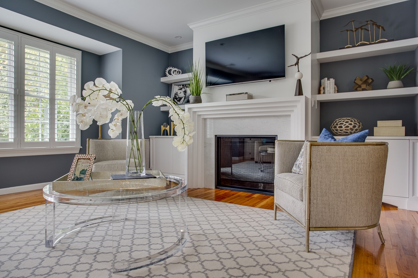 Living room with slate blue walls, white plantation shutters, clear acrylic cocktail table with orchids, and patterned area rug