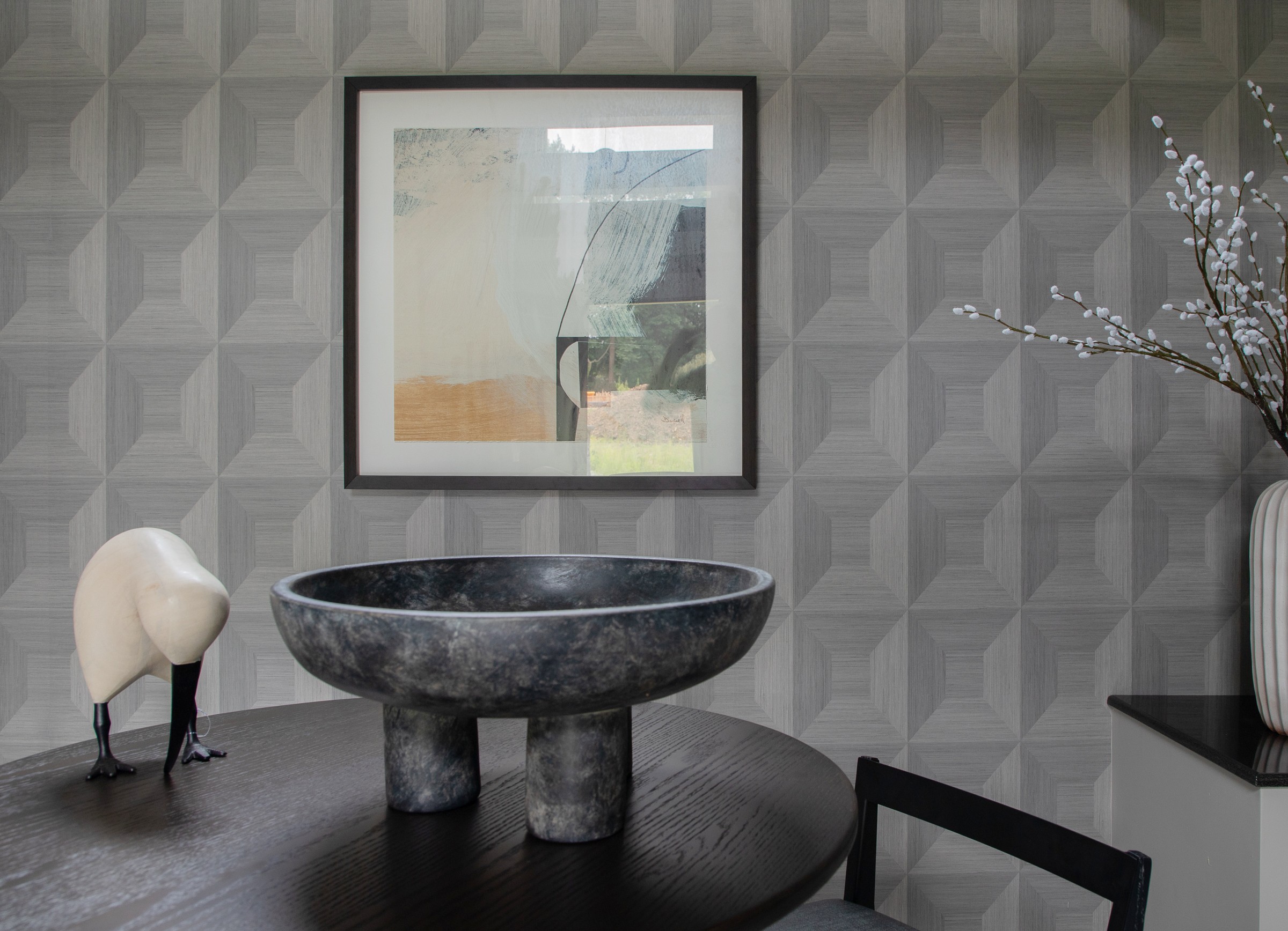 Close-up of dimensional grey geometric wallpaper with framed abstract art, footed stone bowl, and carved bird figurine