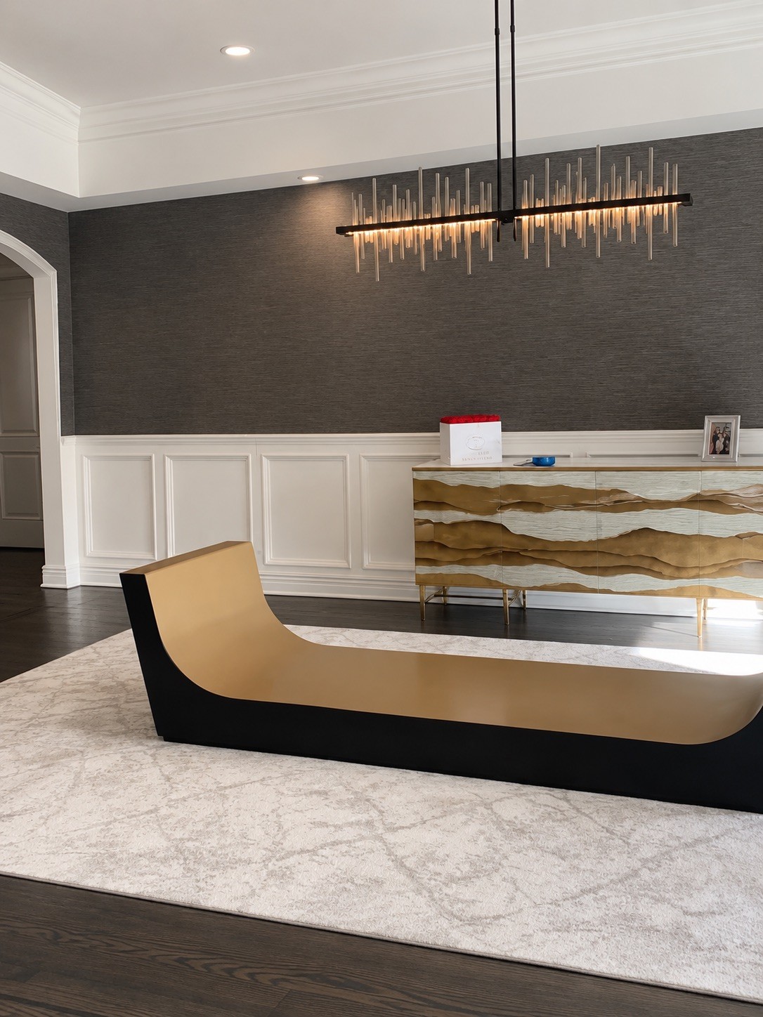 Sculptural antique gold table base installed on a pale area rug before the tabletop's arrival, with a gold-and-white console behind