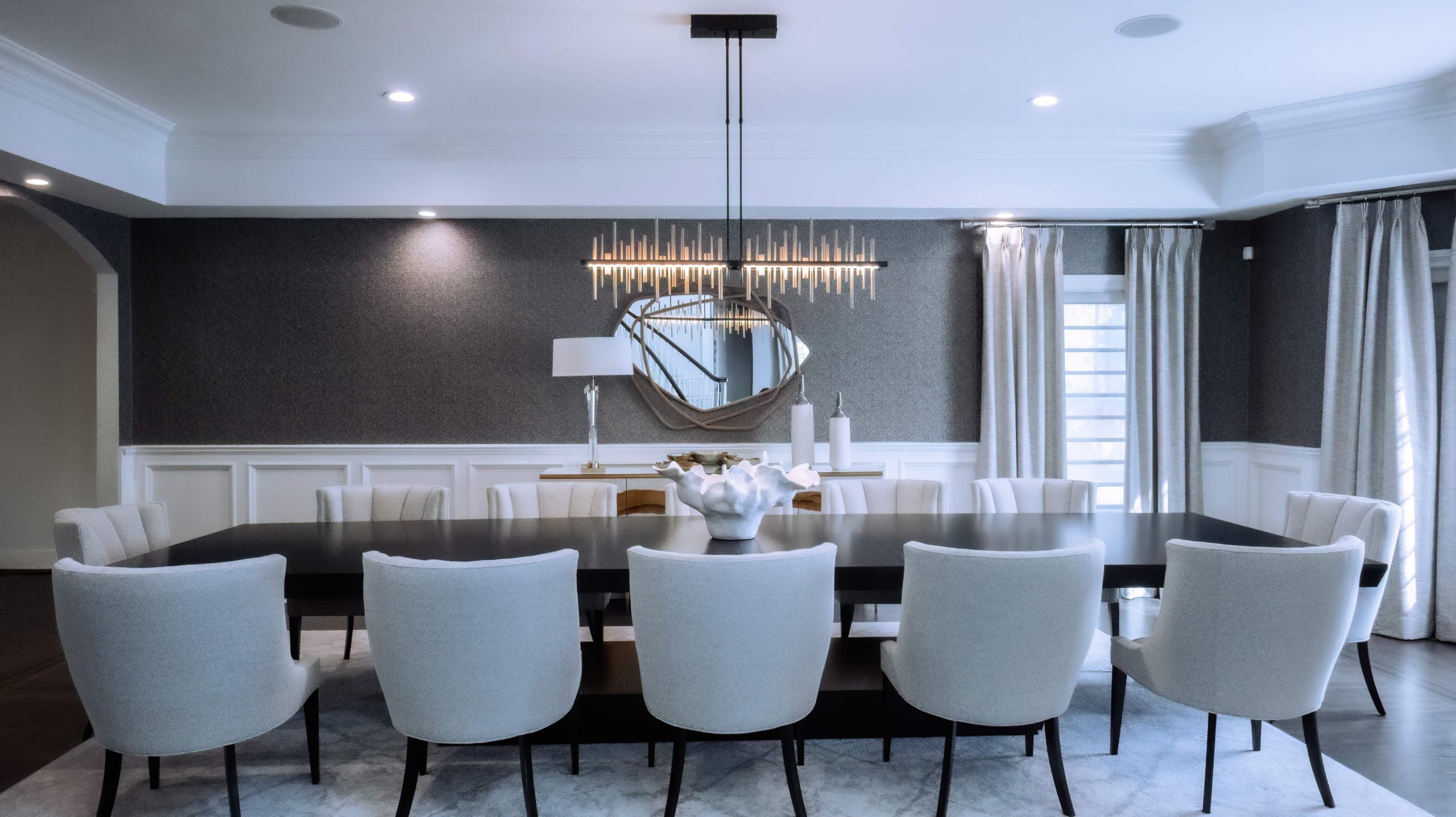 Dining room with charcoal textured wallpaper, sixteen-foot dark wood table, white upholstered chairs, and lit linear chandelier