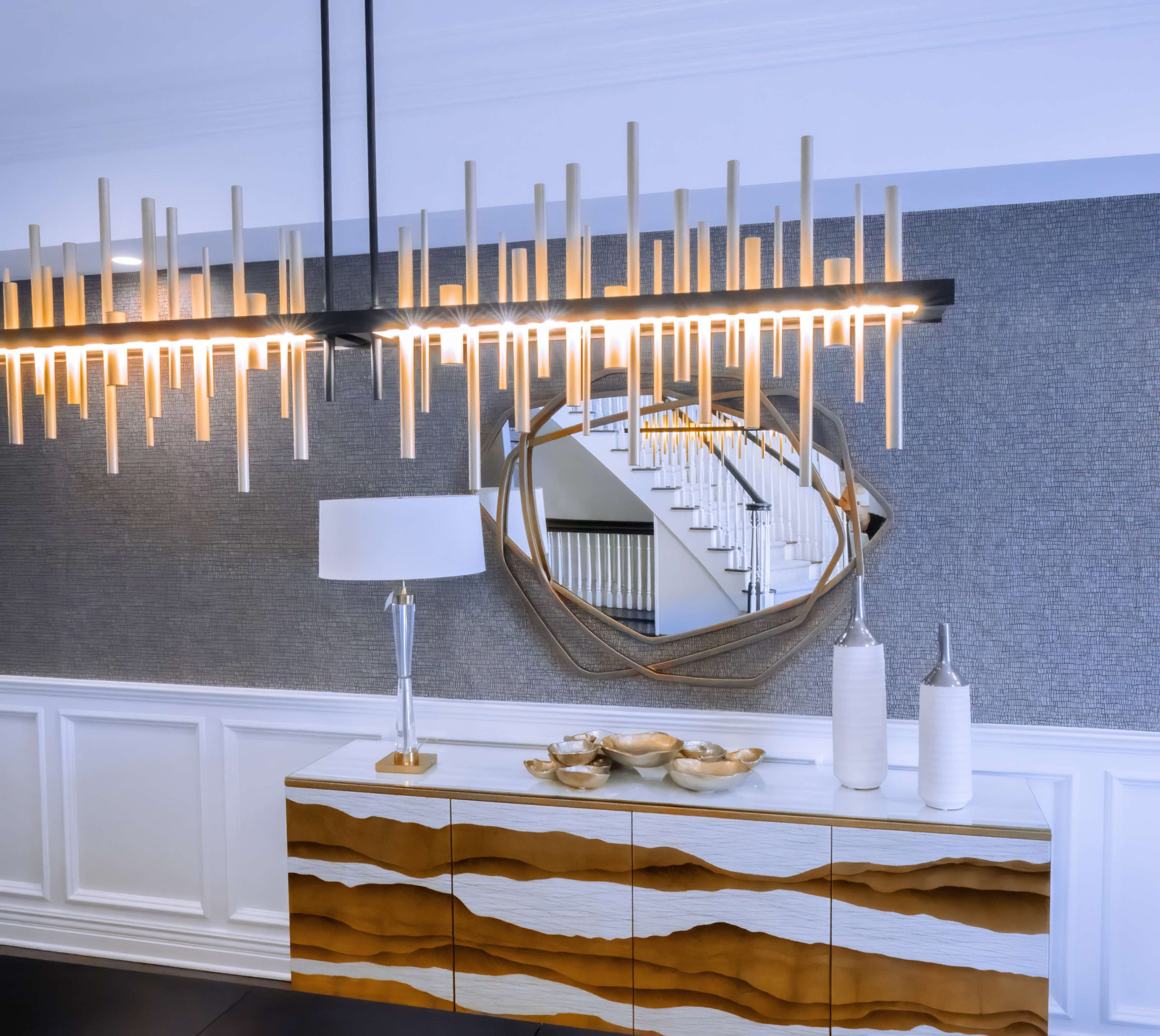 Gold-and-white wave-front console under a geometric mirror and lit linear chandelier against textured charcoal wallpaper